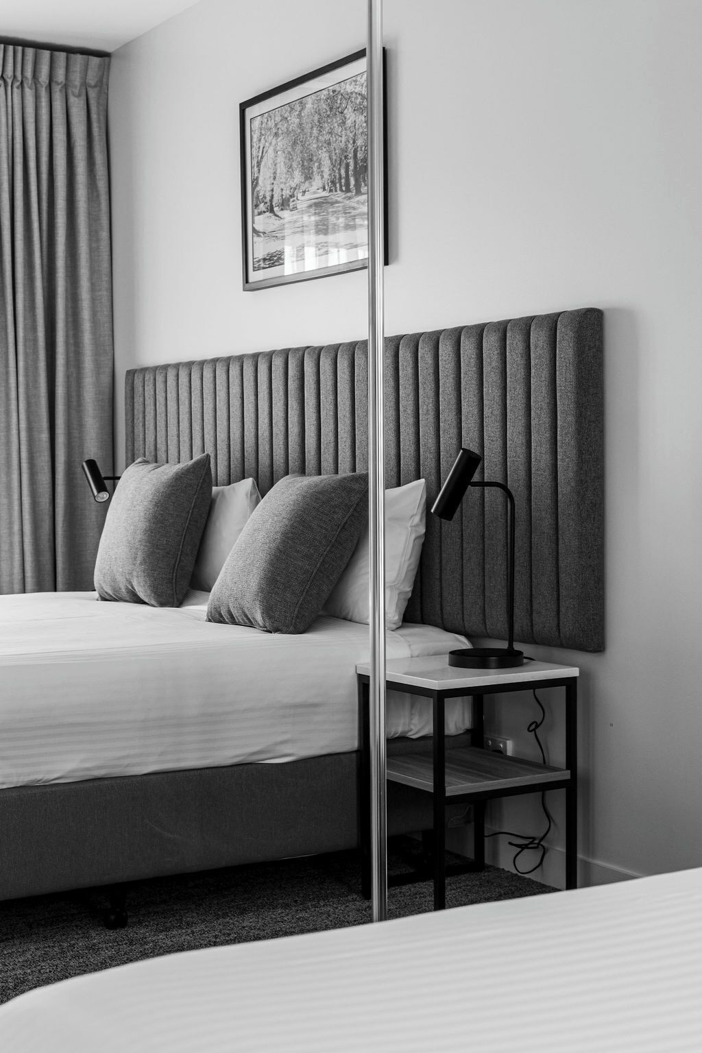 Una habitación de hotel moderna con una cama grande, un cabecero con líneas verticales, dos lámparas de noche y un cuadro enmarcado en la pared.