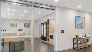 A modern reception area with a glass door, a reception desk labeled 'QUEST,' a wooden shelving unit, and a framed picture on the wall.
