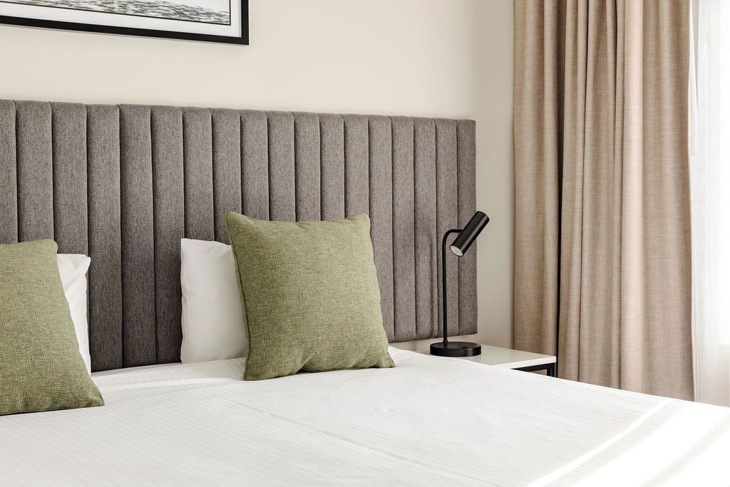 A hotel room with a large bed, a grey headboard, and a bedside table with a lamp.