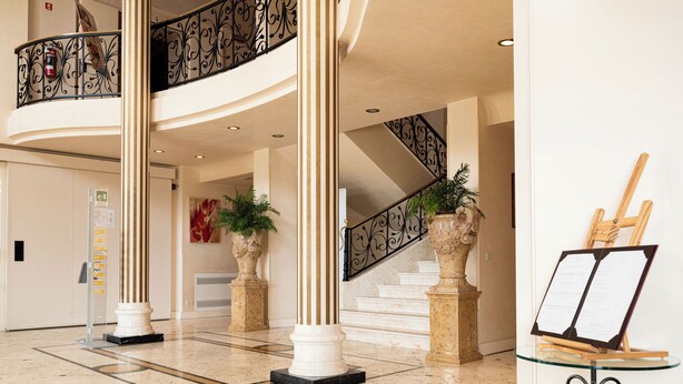 Un vestíbulo con piso de mármol, columnas blancas, una escalera con barandillas negras y plantas en macetas.