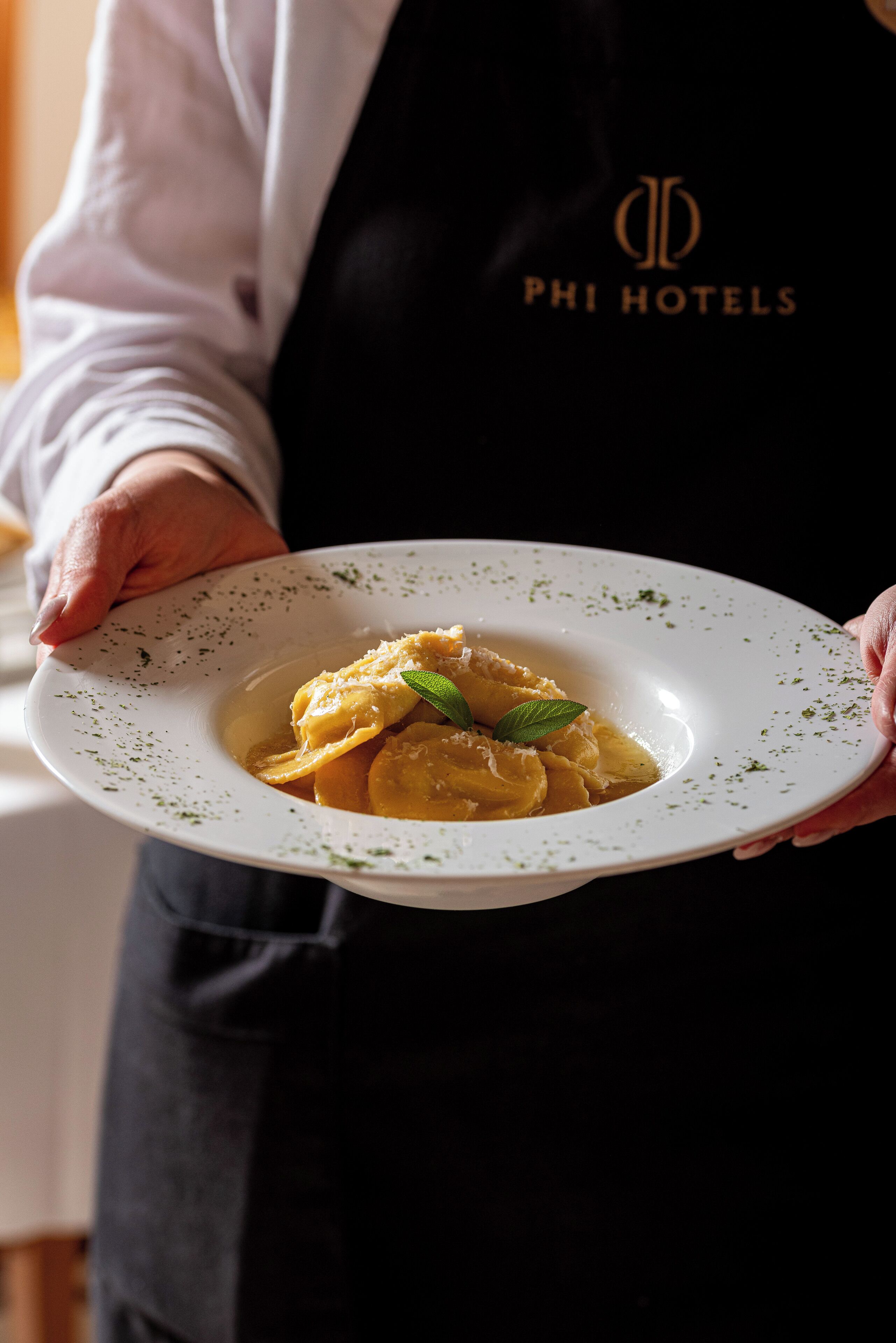 Eine Person hält einen weißen Teller mit Ravioli und einer grünen Blattgarnitur. Die Person trägt eine schwarze Schürze mit dem Aufdruck "PHI HOTELS".