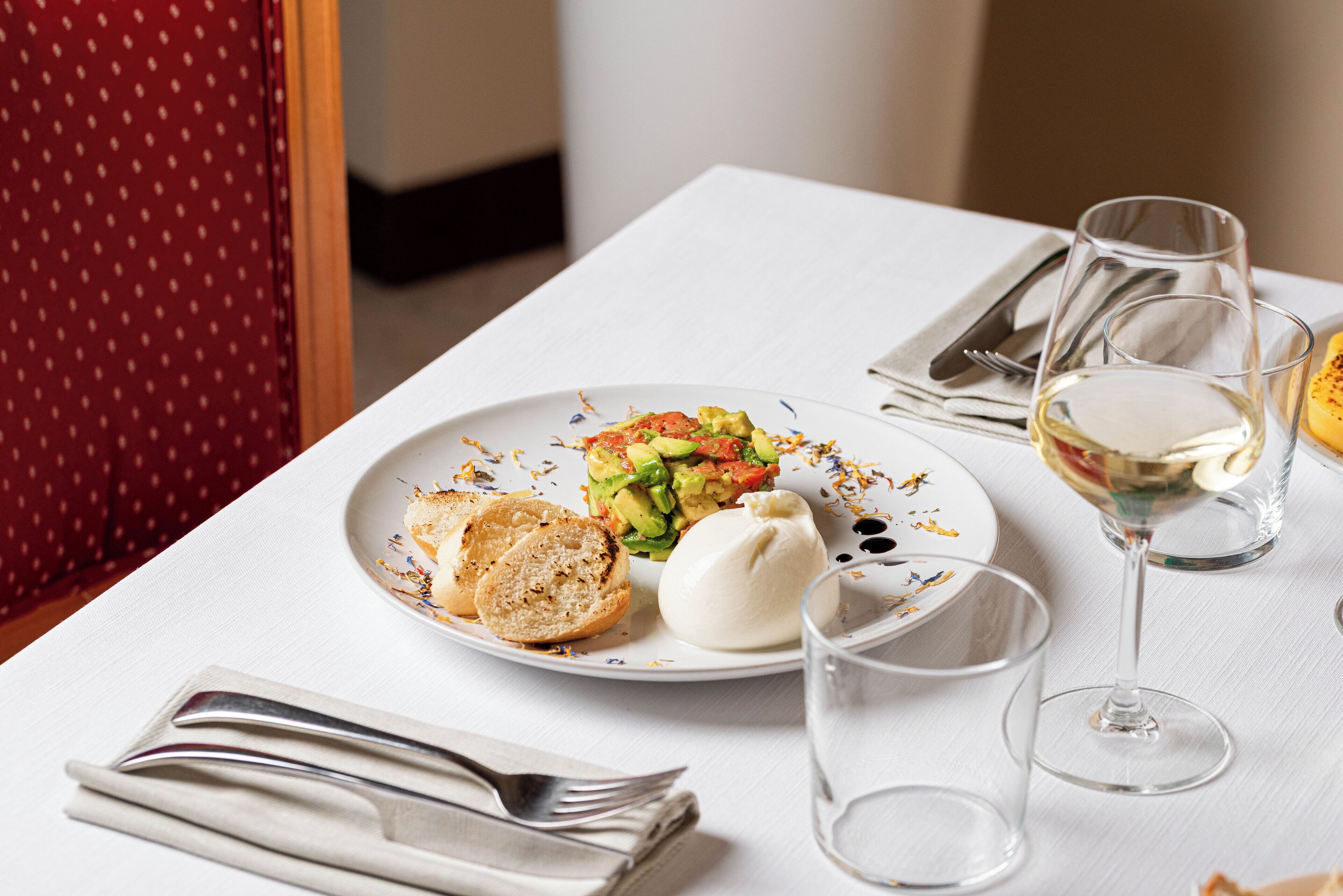 Una mesa de comedor con un plato de comida, una copa de vino blanco y cubiertos.