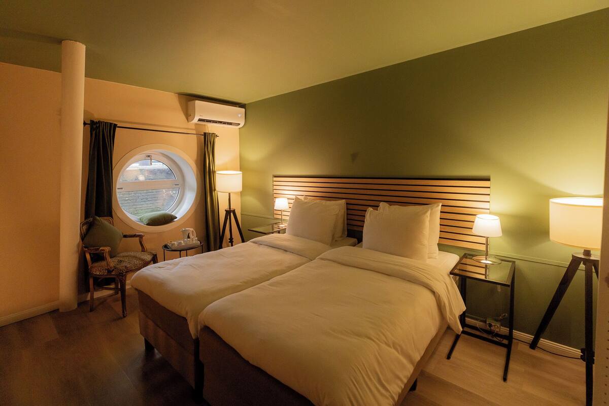 A hotel room with a large bed, two bedside tables with lamps, a chair, a round window, and an air conditioning unit.
