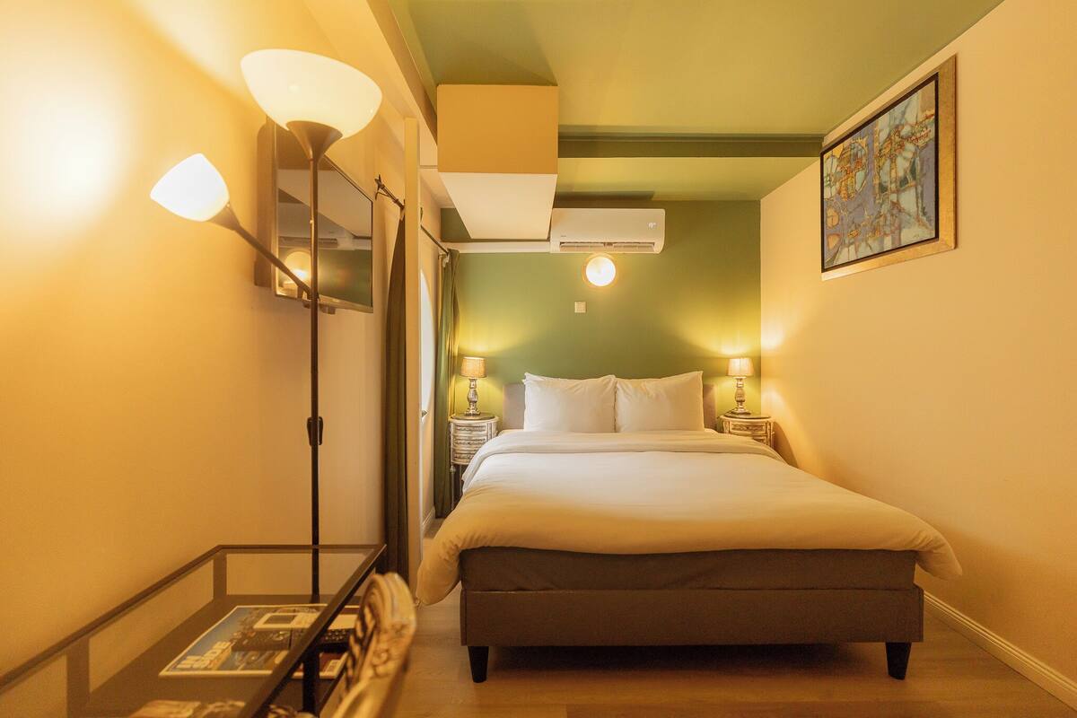 A hotel room with a bed, bedside lamps, an air conditioner, a painting on the wall, and a glass table with magazines.