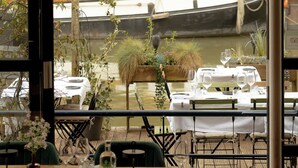 Un restaurant avec des tables dressées pour le service, une vue sur les bateaux sur l’eau et des plantes d’intérieur.