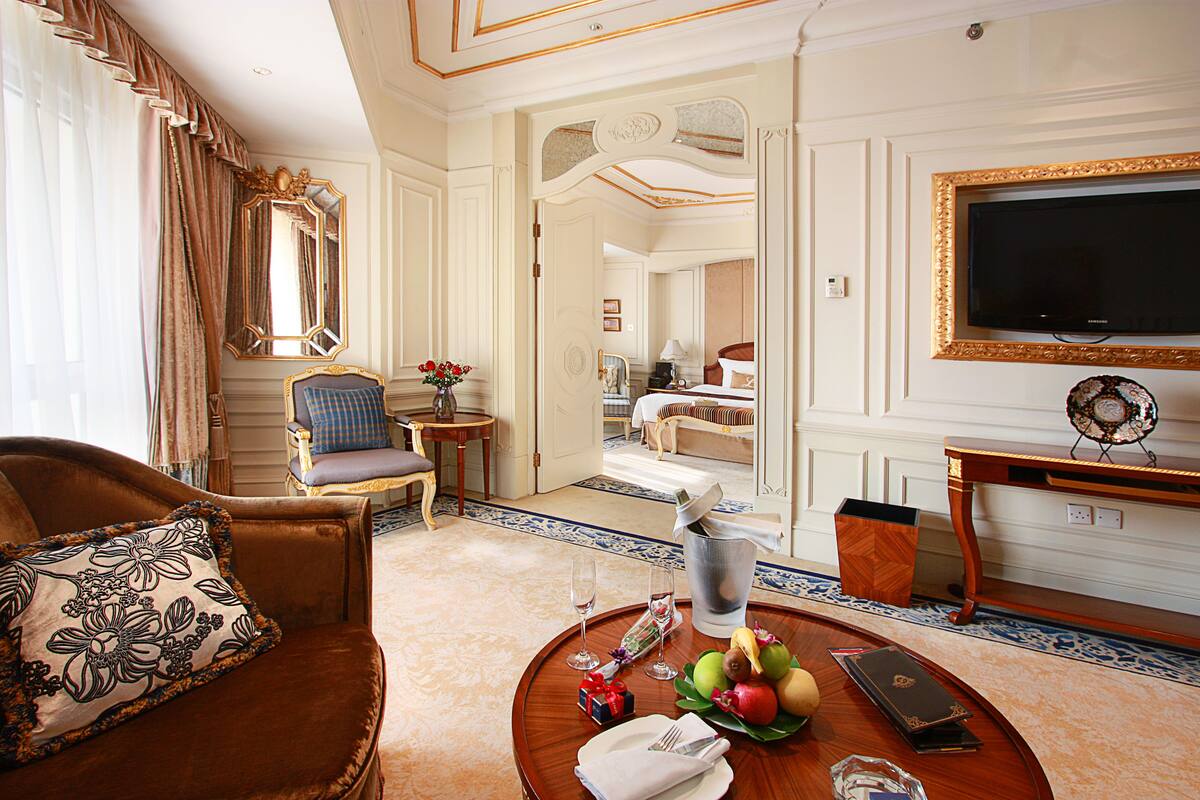 Habitación de hotel con sofá de cuero marrón, una mesa redonda con frutas y un libro, televisor en la pared y un espejo con marco dorado.