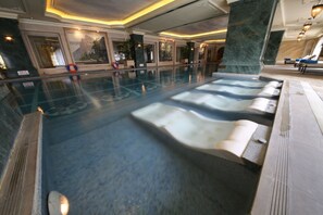 A large indoor swimming pool with multiple hot tubs, surrounded by marble columns and framed artwork.