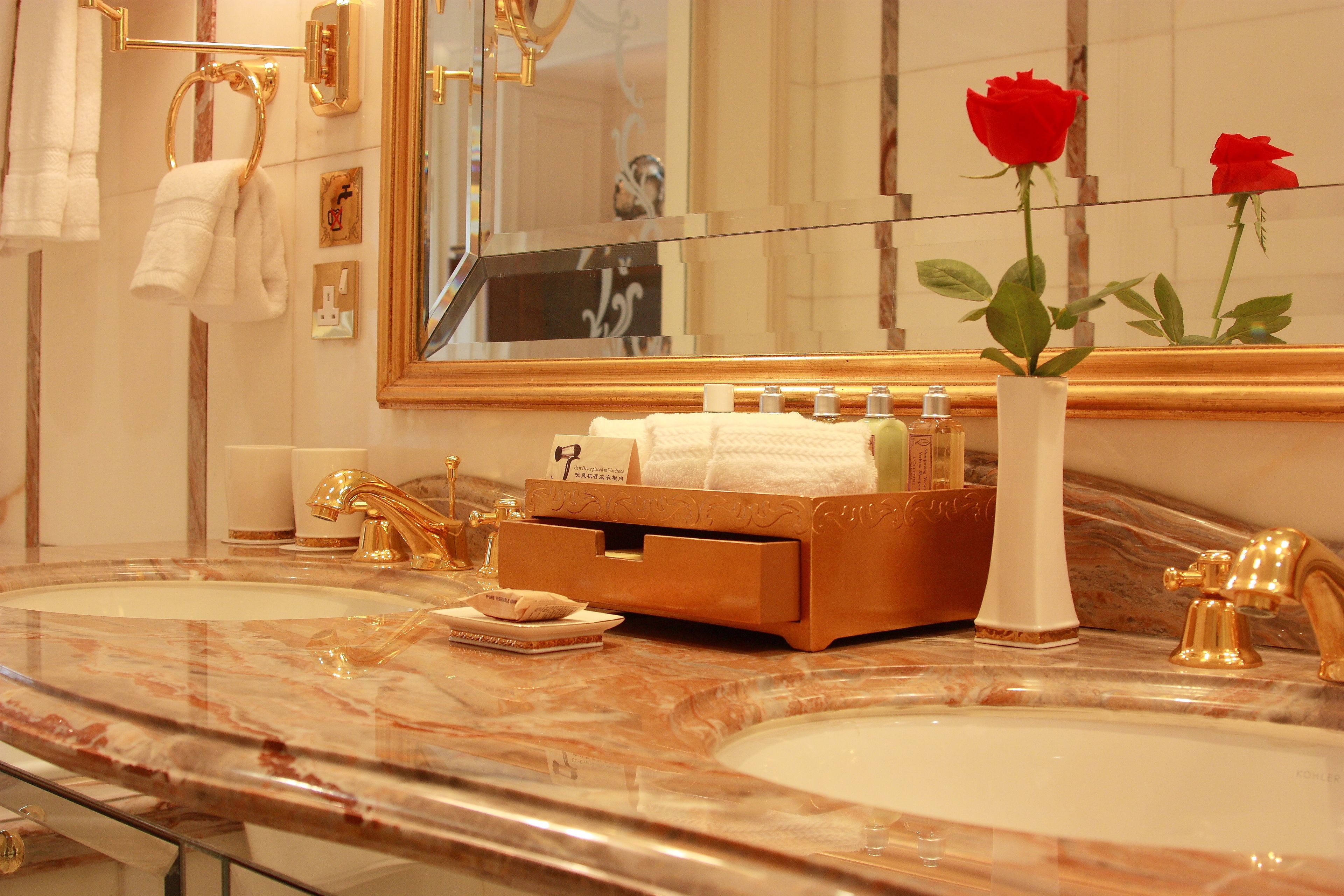 A bathroom with two sinks, gold faucets, a marble countertop, a vanity with a mirror, and a vase with red roses.