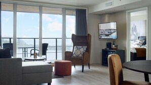 A modern hotel room with a large window offering a view of the water, a comfortable seating area with a sofa and armchairs, a flat-screen TV mounted on the wall, and a small side table with a lamp.