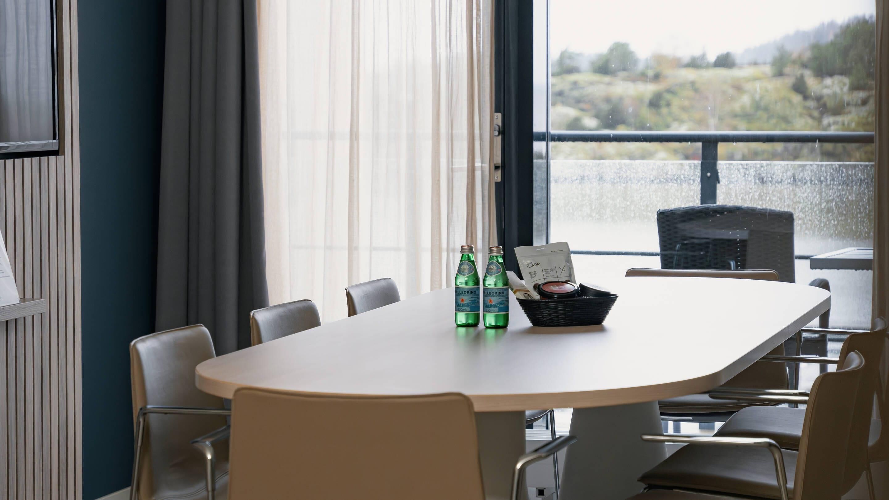 A conference room with a white table, six chairs, two bottles of water, a basket with a coffee cup, and a view of a landscape through large windows.