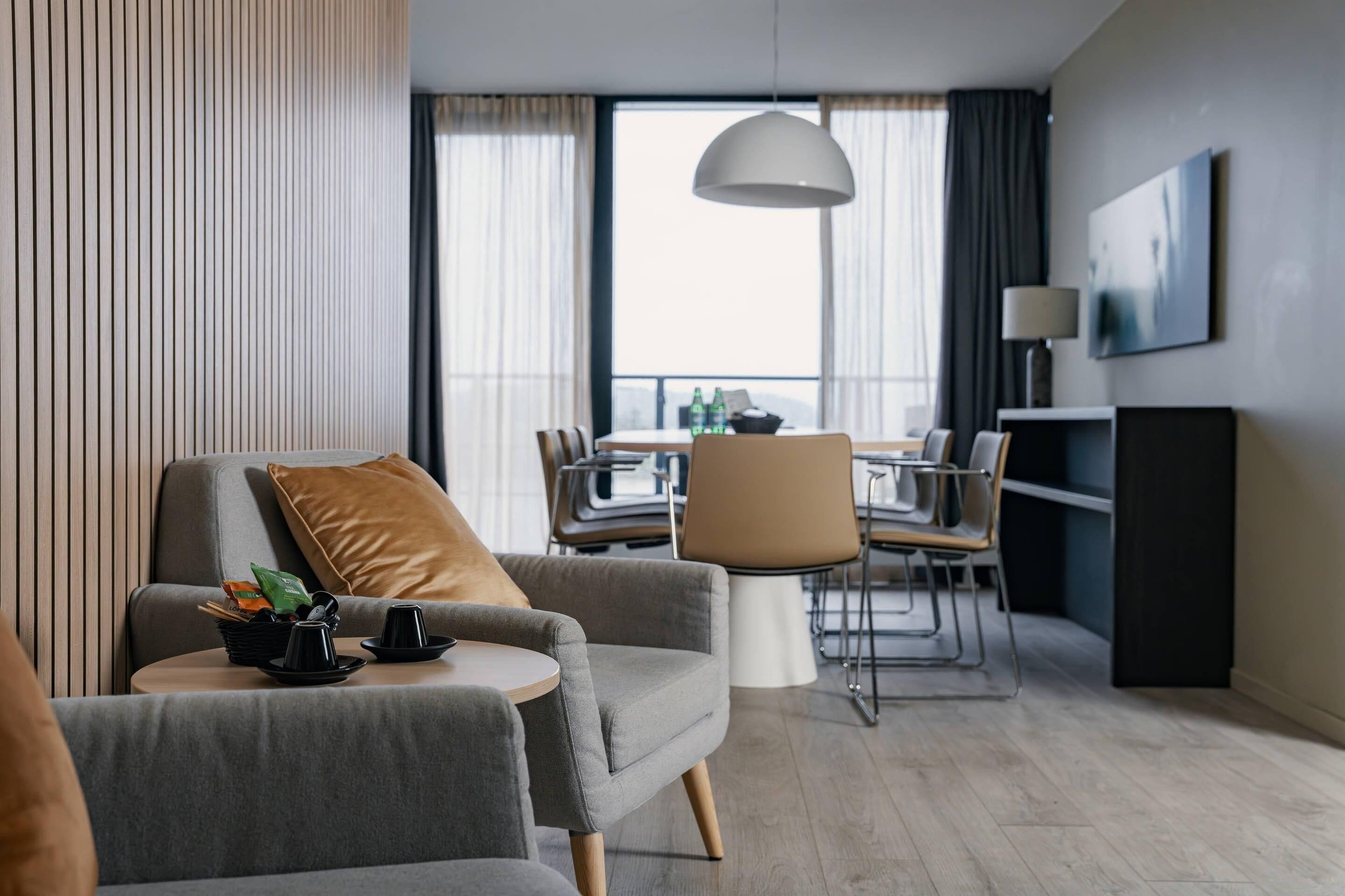 A modern hotel room with a grey sofa, a round coffee table, a dining area with a table and chairs, and a large window with curtains.
