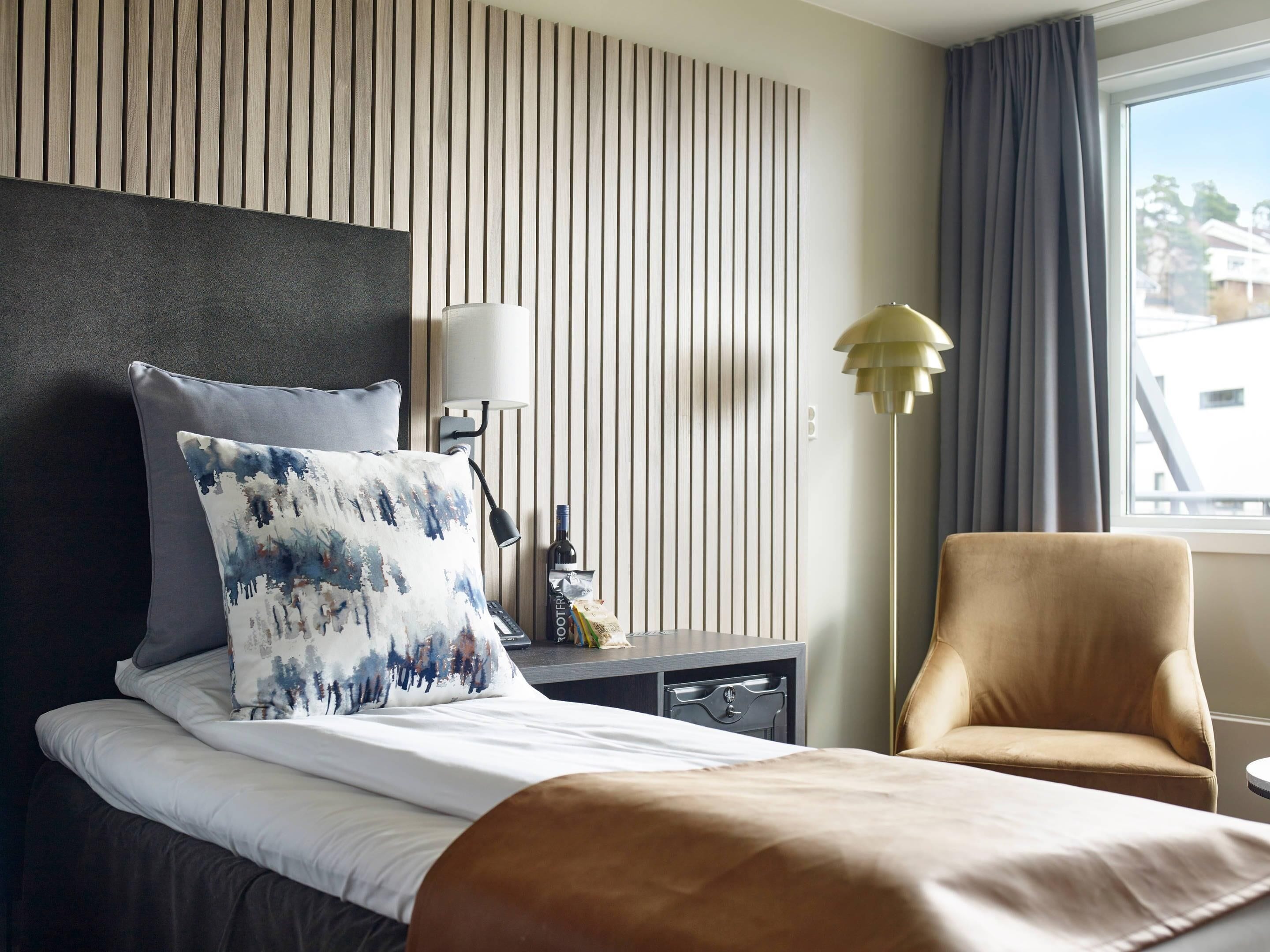 Una habitación de hotel con cama, mesita de noche, lámpara, silla y ventana con cortinas.