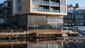 A modern multi-story building with large windows and balconies, situated by a calm body of water.