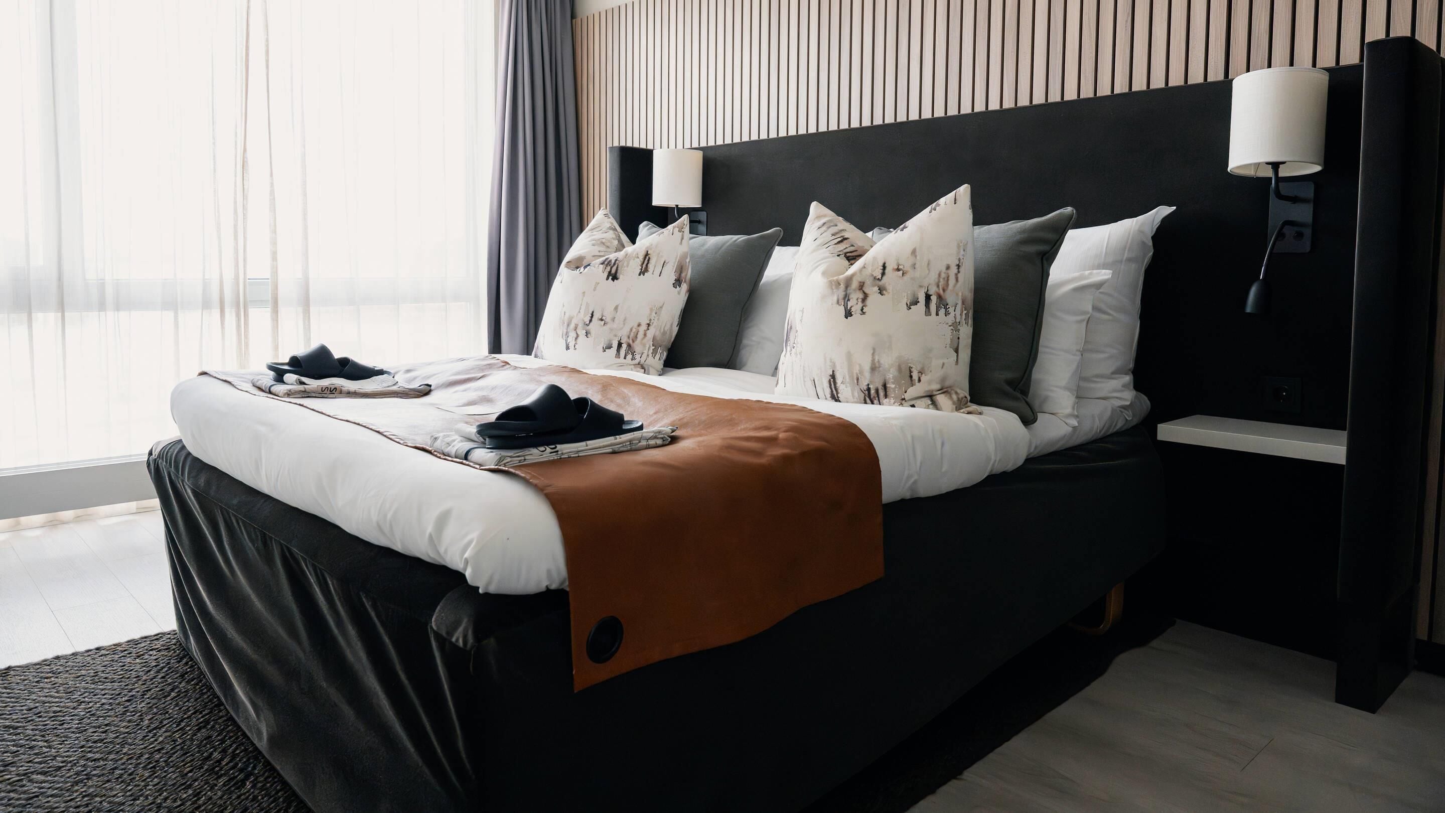 A modern hotel room with a large bed, a bedside table with a lamp, and a view of the city through sheer curtains.