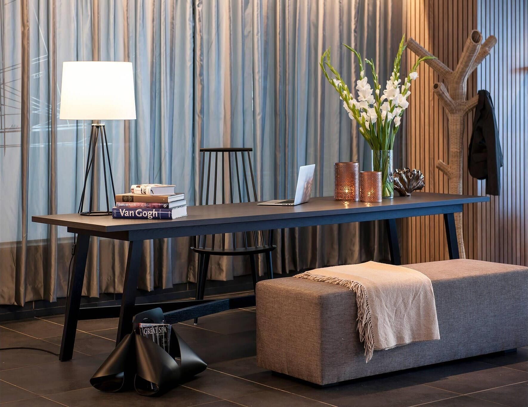 A modern hotel room with a dark wooden table, a laptop, a stack of books, a lamp, and a decorative vase with flowers.