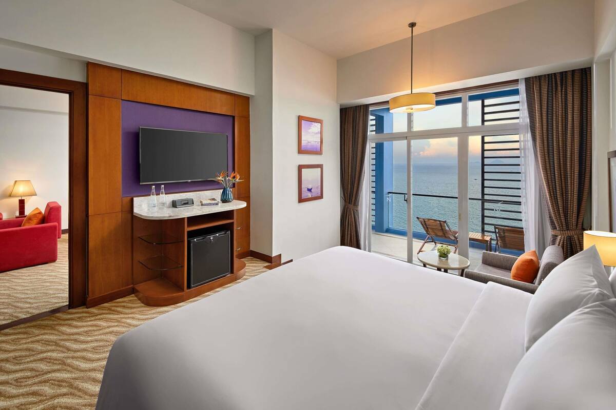 A hotel room with a bed, a TV, a desk with a computer, a small table, and a view of the sea.