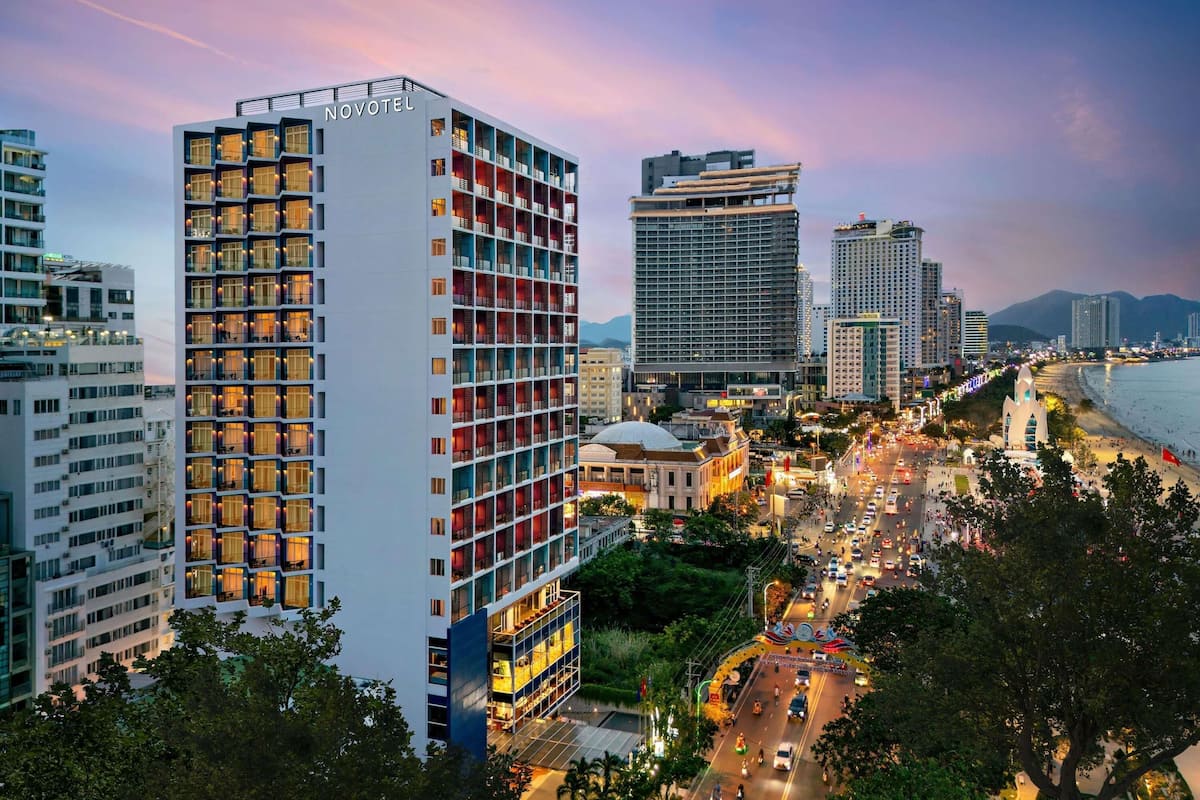 A modern hotel with multiple floors, a prominent 'NOVOTEL' sign, and a view of a busy street and beach in the background.