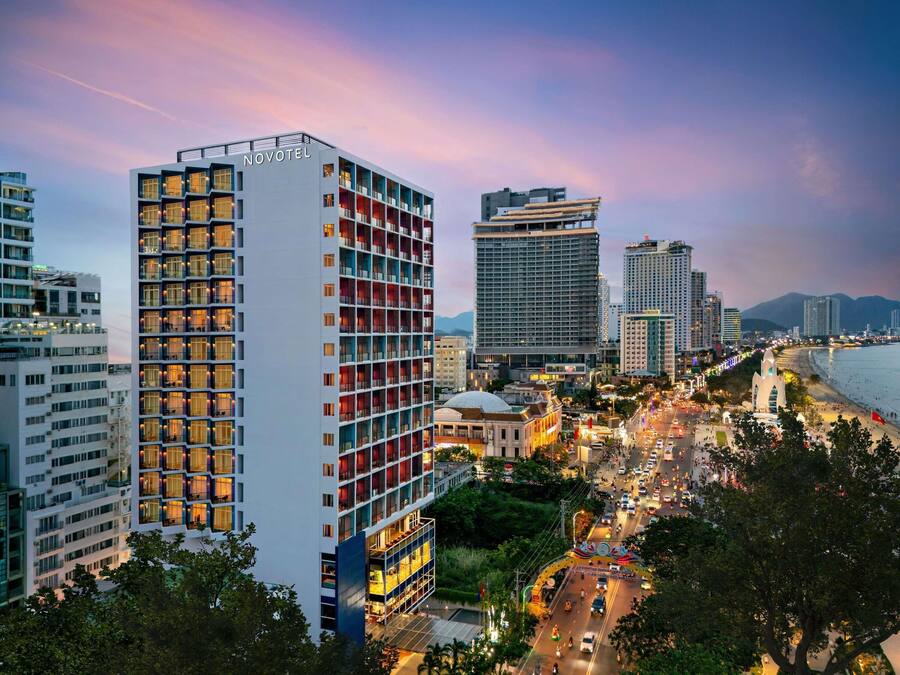 Un hotel moderno con varios pisos, un llamativo letrero de 'NOVOTEL', y una vista de una calle concurrida y una playa al fondo.