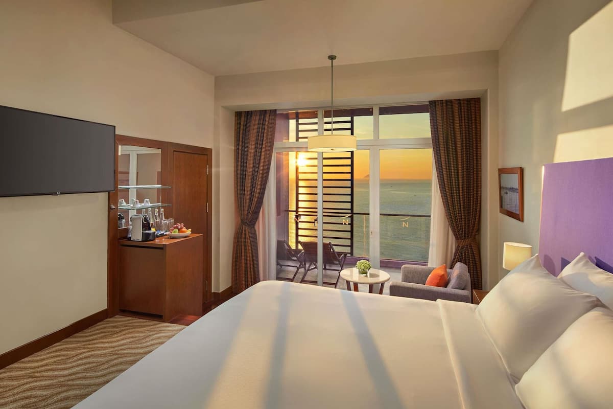 A hotel room with a bed, a desk with a coffee machine, a small table, and a balcony with a view of the sea.