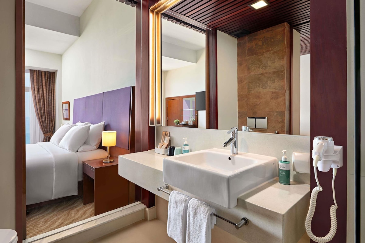 Habitación de hotel con cama, mesita de noche con lámpara, un espejo grande, y baño con lavamanos y toallero.