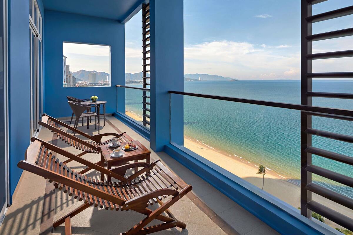 A balcony with wooden chairs and a table, overlooking a beach and ocean.