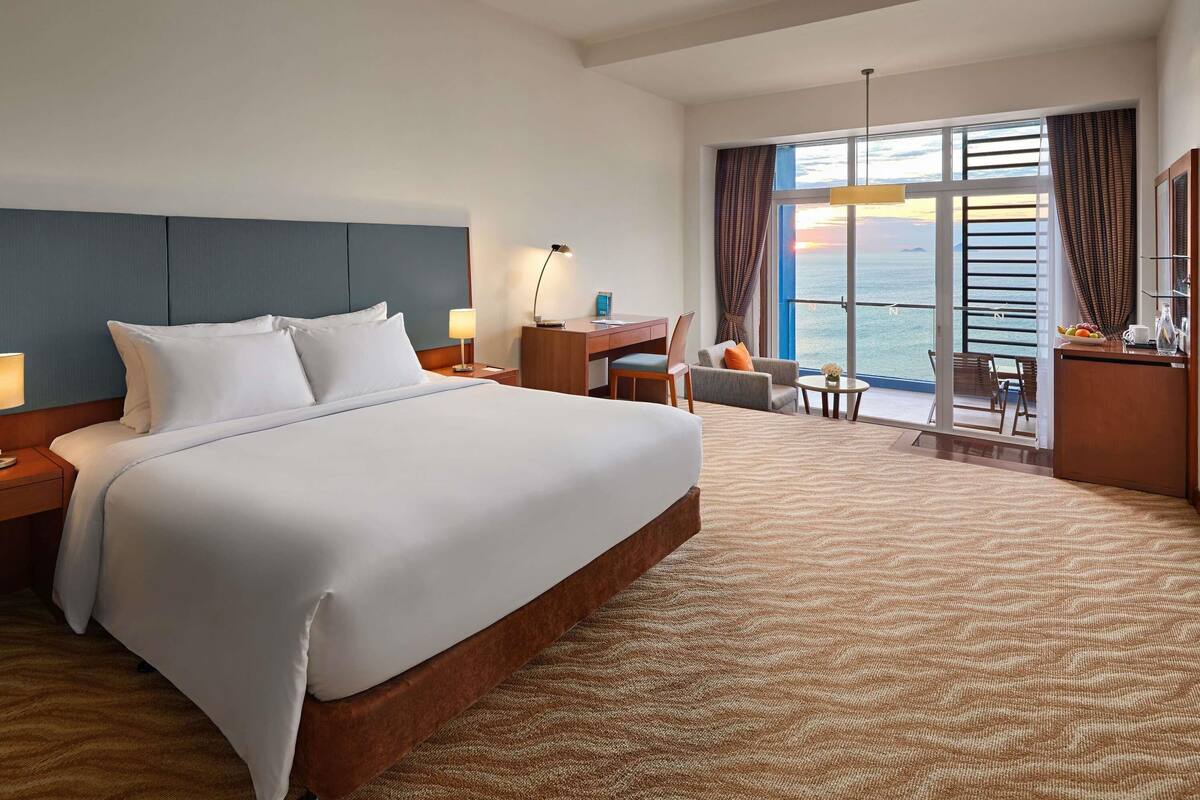 A hotel room with a large bed, a desk, a chair, and a view of the sea through a large window.