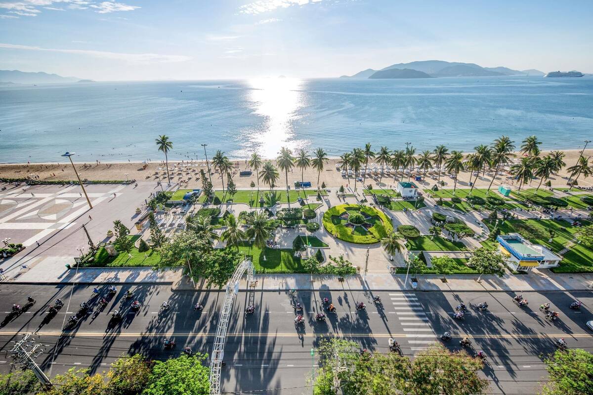 An aerial view of a coastal area with a beach, palm trees, and a road with motorcyclists.