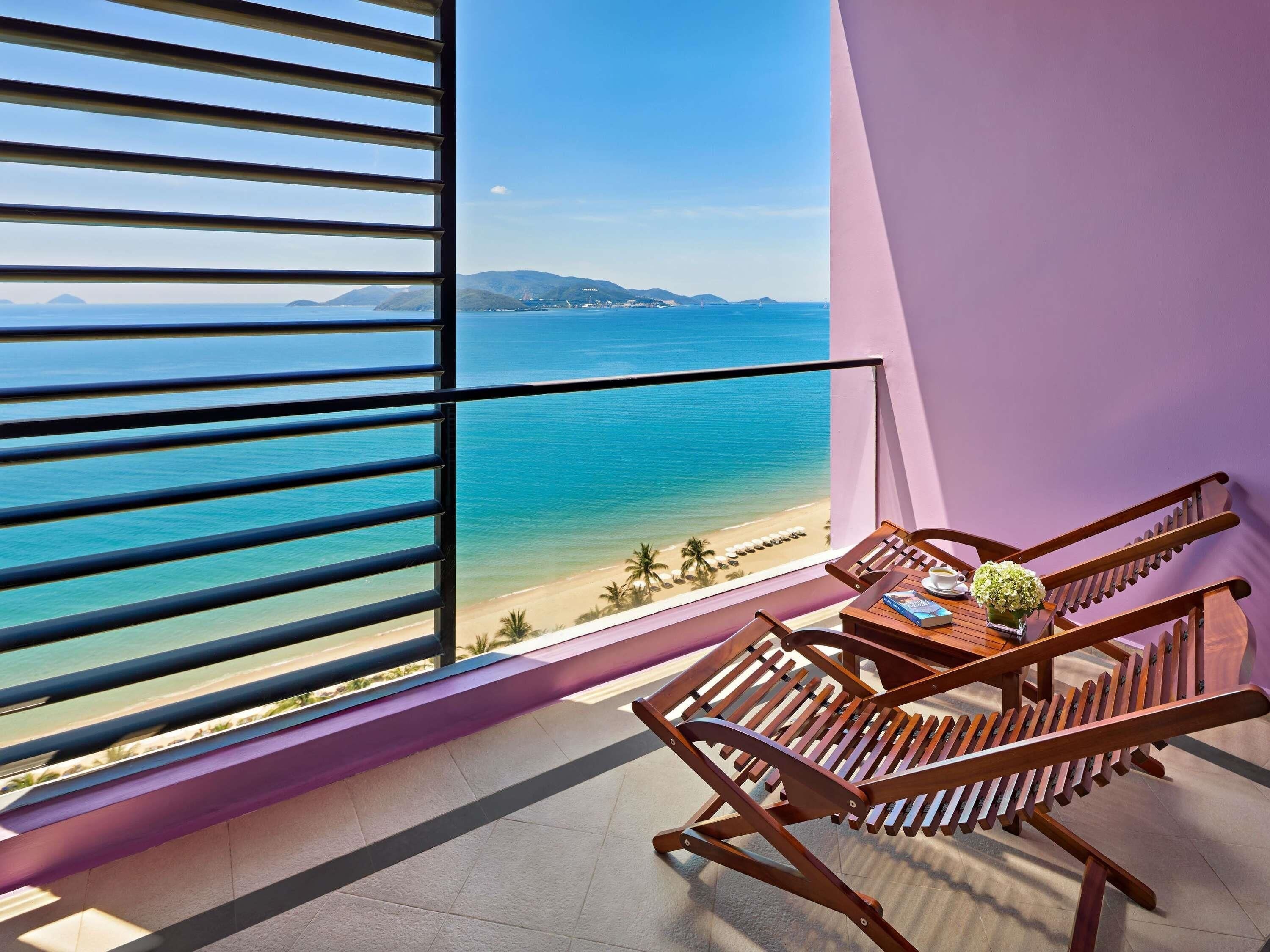 A balcony with a view of the ocean, a wooden chair, and a table with a book and a flower vase.