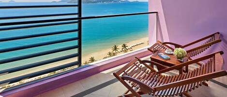 A balcony with a view of the ocean, a wooden chair, and a table with a book and a flower vase.