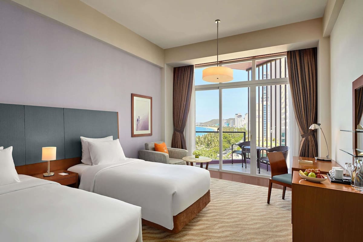 A hotel room with two beds, a desk, a chair, a small table, and a balcony with a view.