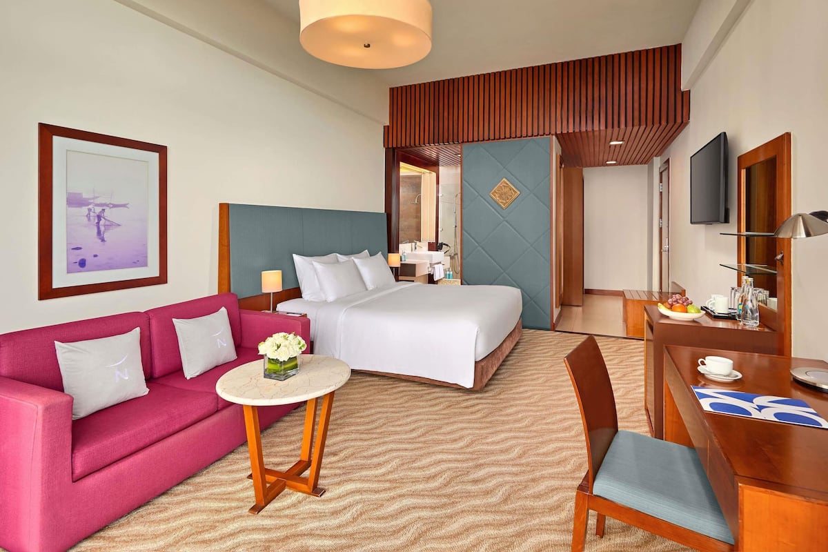 A hotel room with a bed, a pink sofa, a small round table with a vase, a wooden desk with a chair, and a TV on the wall.