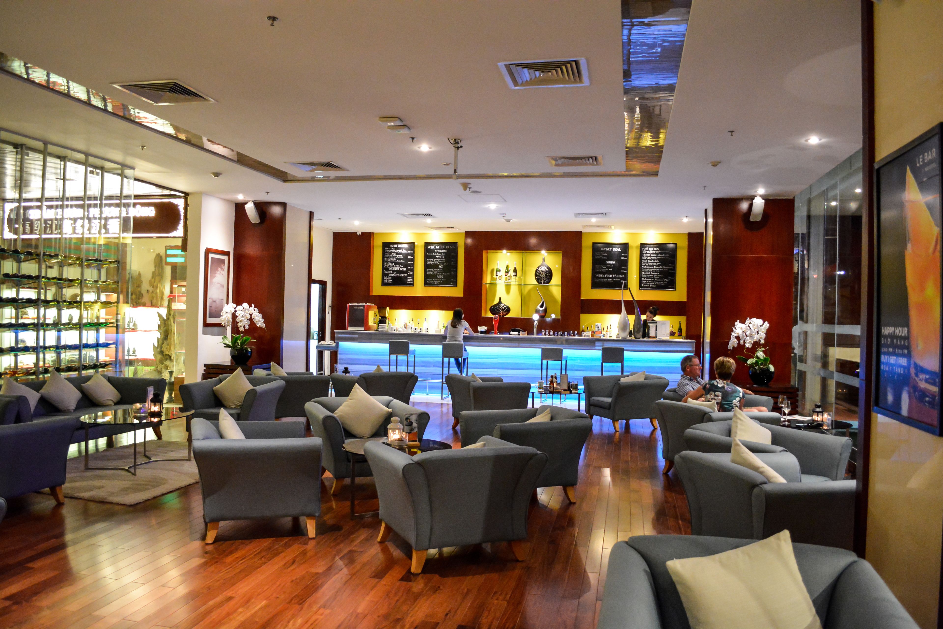 Un vestíbulo de hotel con sofás grises, una zona de bar y una vitrina de cristal con objetos.