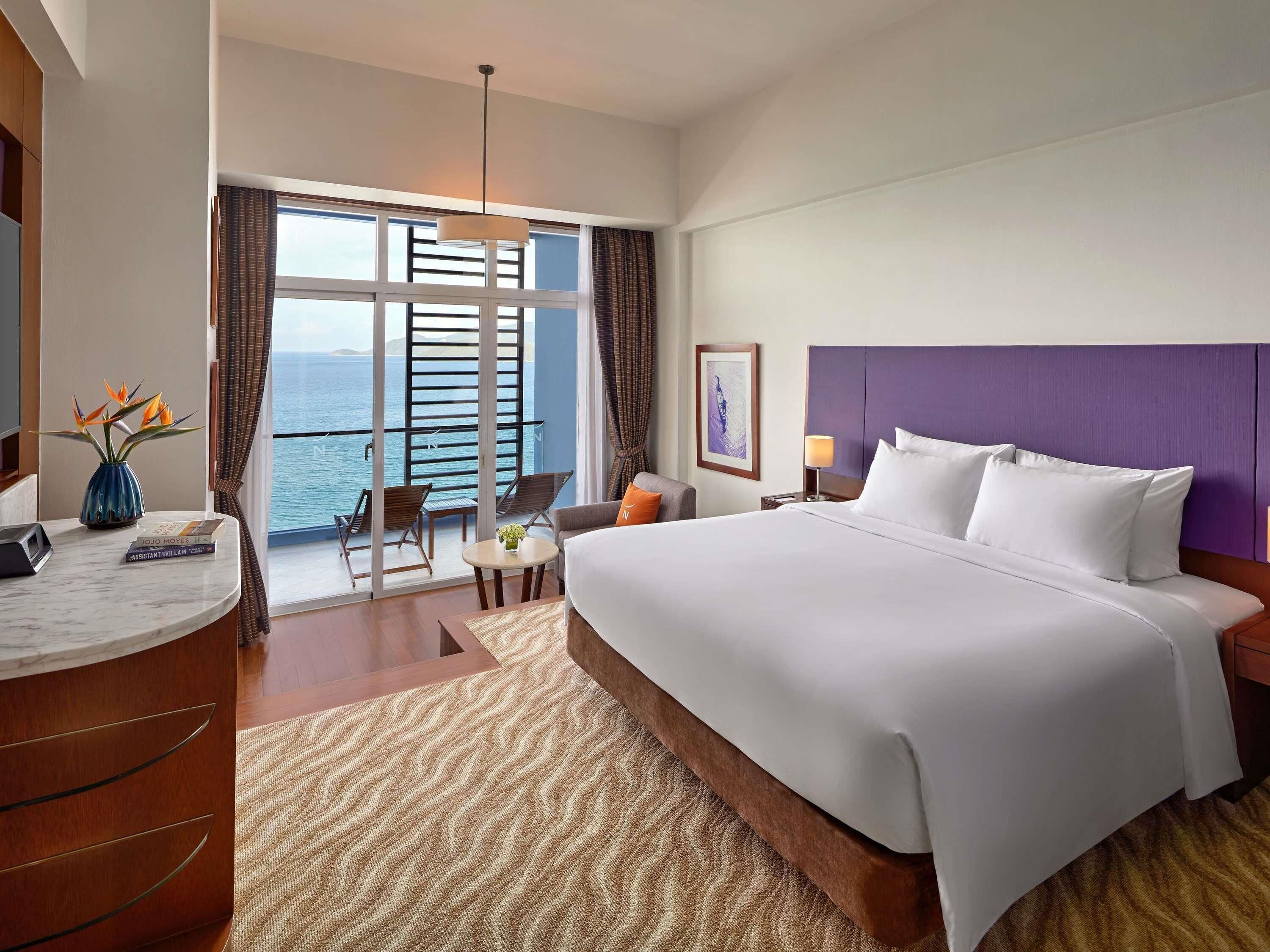 A hotel room with a large bed, a desk with a chair, a small table with a vase, a view of the sea, and a balcony with chairs.