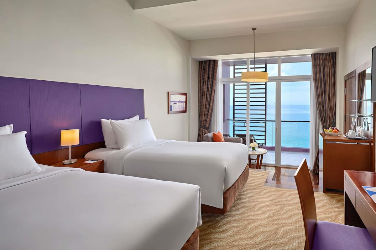A hotel room with two beds, a desk, a chair, a lamp, a window with curtains, and a view of the sea.
