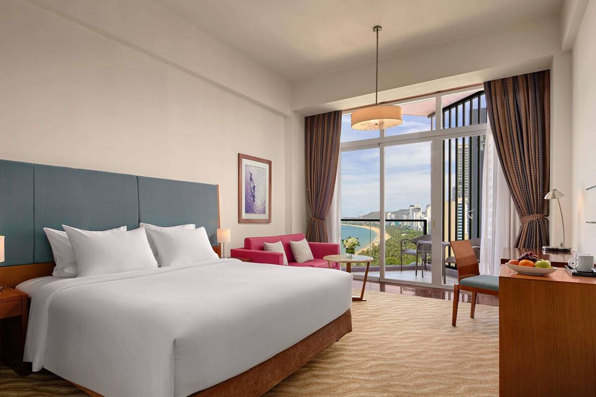 A hotel room with a large bed, a desk with a lamp, a chair, a small table with fruit, and a view of the city through large windows.
