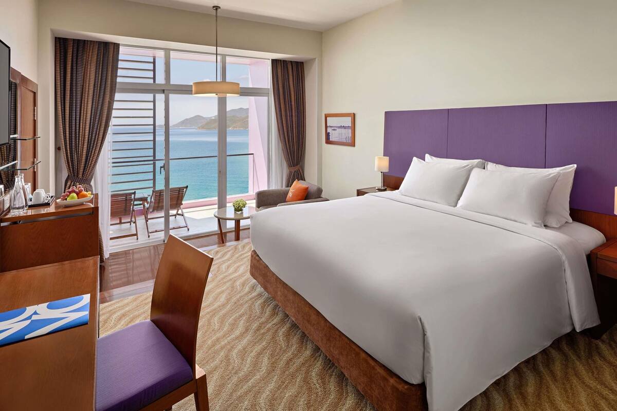 A hotel room with a large bed, a desk with a chair, a small table with a lamp, and a view of the sea.