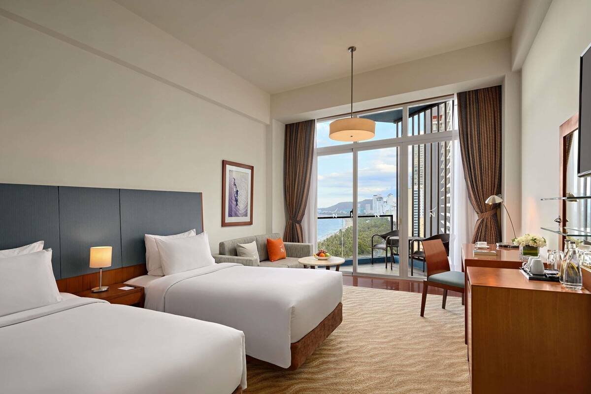 A hotel room with two beds, a desk, a chair, a large window with a view, and a ceiling light.