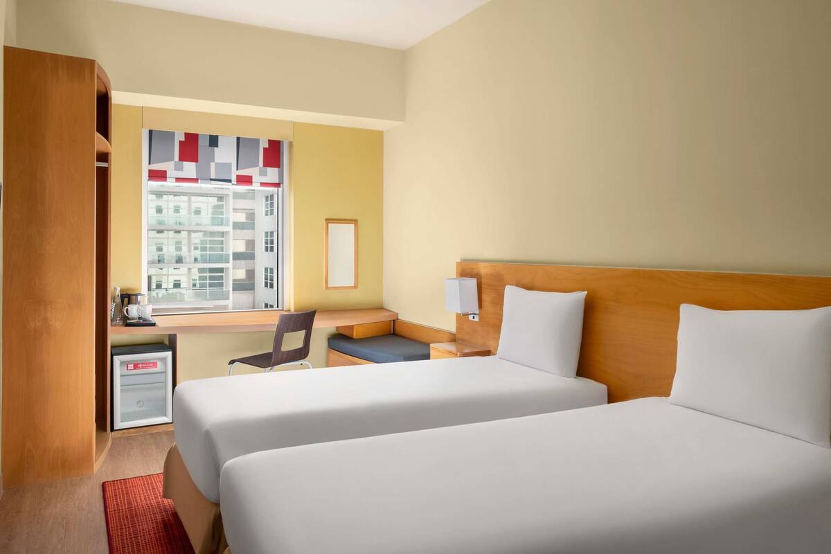 A hotel room with two beds, a desk, a chair, a small table, a window with a view of buildings, and a red rug.