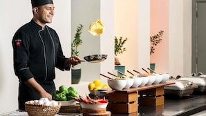 Un chef vêtu d’un uniforme noir et coiffé d’une toque de chef, tenant une poêle contenant une crêpe, se tient dans une cuisine moderne où est installé un buffet.