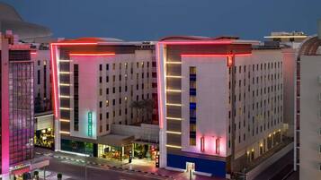 Un hôtel à plusieurs étages doté d'une architecture moderne, avec une entrée imposante marquée par un panneau néon rouge, un espace de stationnement à l'avant et une vue dégagée sur le paysage urbain environnant.