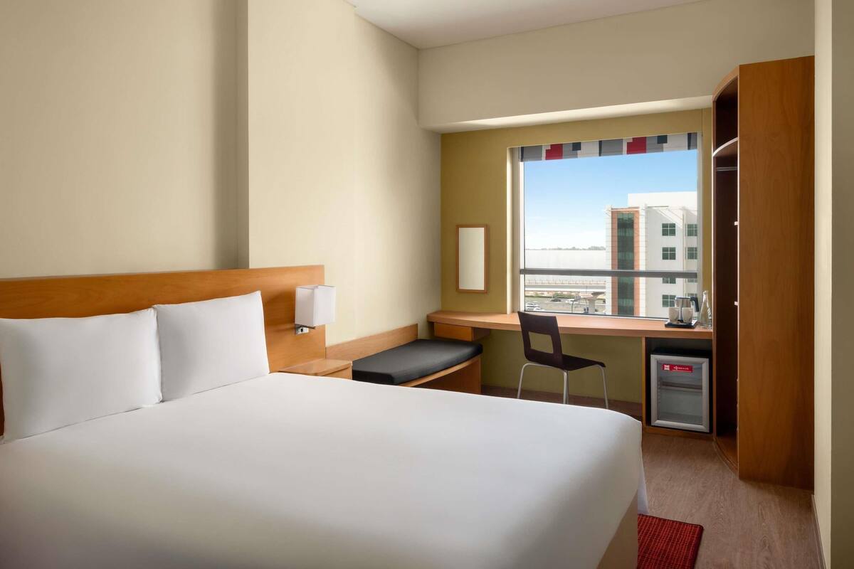 A hotel room with a bed, a desk, a chair, a small fridge, and a window with a view of buildings.