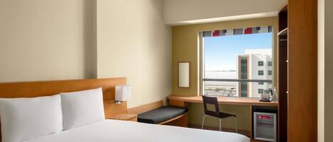 Habitación de hotel con cama, escritorio, silla, refrigerador pequeño y ventana con vista a edificios.