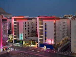 Un hotel de varios pisos con diseño moderno, que destaca por una entrada con un letrero de neón rojo, múltiples balcones y un área de estacionamiento al frente.