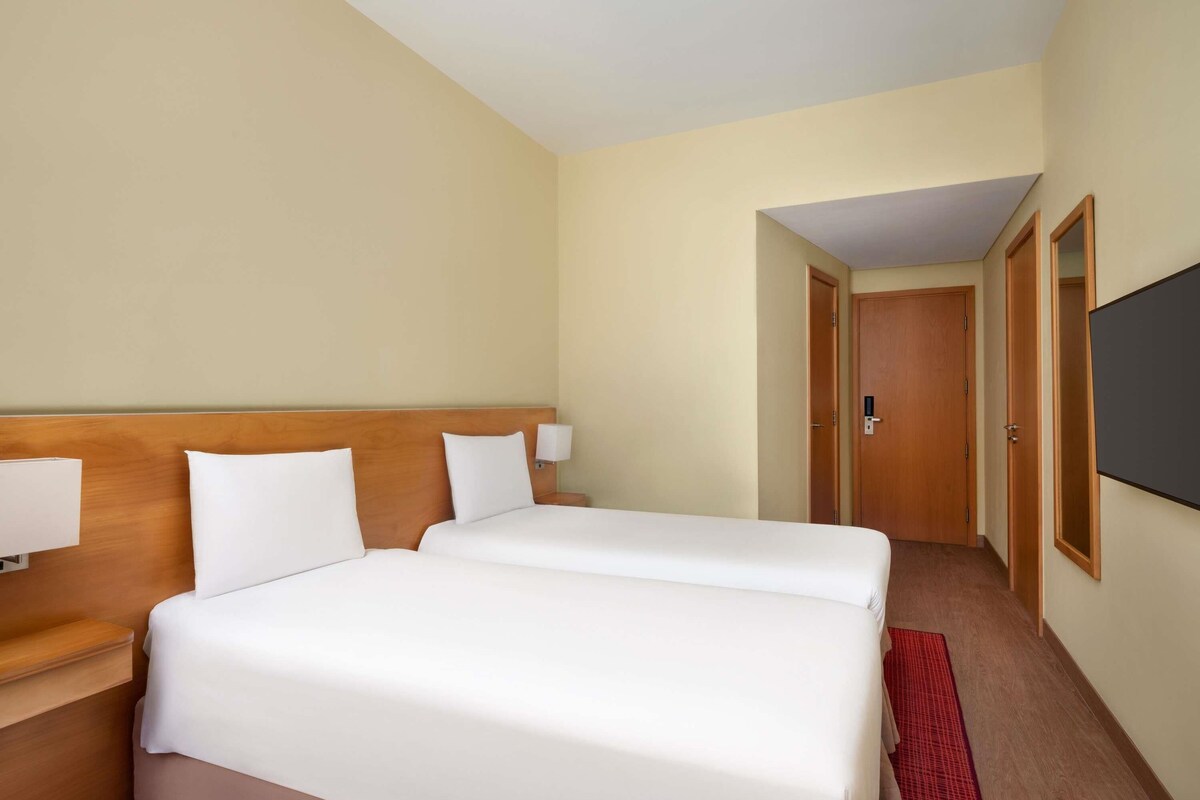 Habitación de hotel con dos camas, cabecera de madera, lámparas de noche, televisor y alfombra roja.