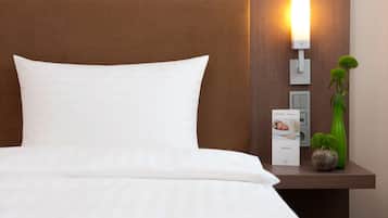 A neatly made bed with white linens, a brown headboard, a bedside table with a lamp, a small potted plant, and a brochure.