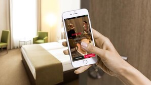 Una mano sosteniendo un teléfono inteligente que muestra una interfaz de reserva de hotel, con una cama y una planta de habitación visibles al fondo.