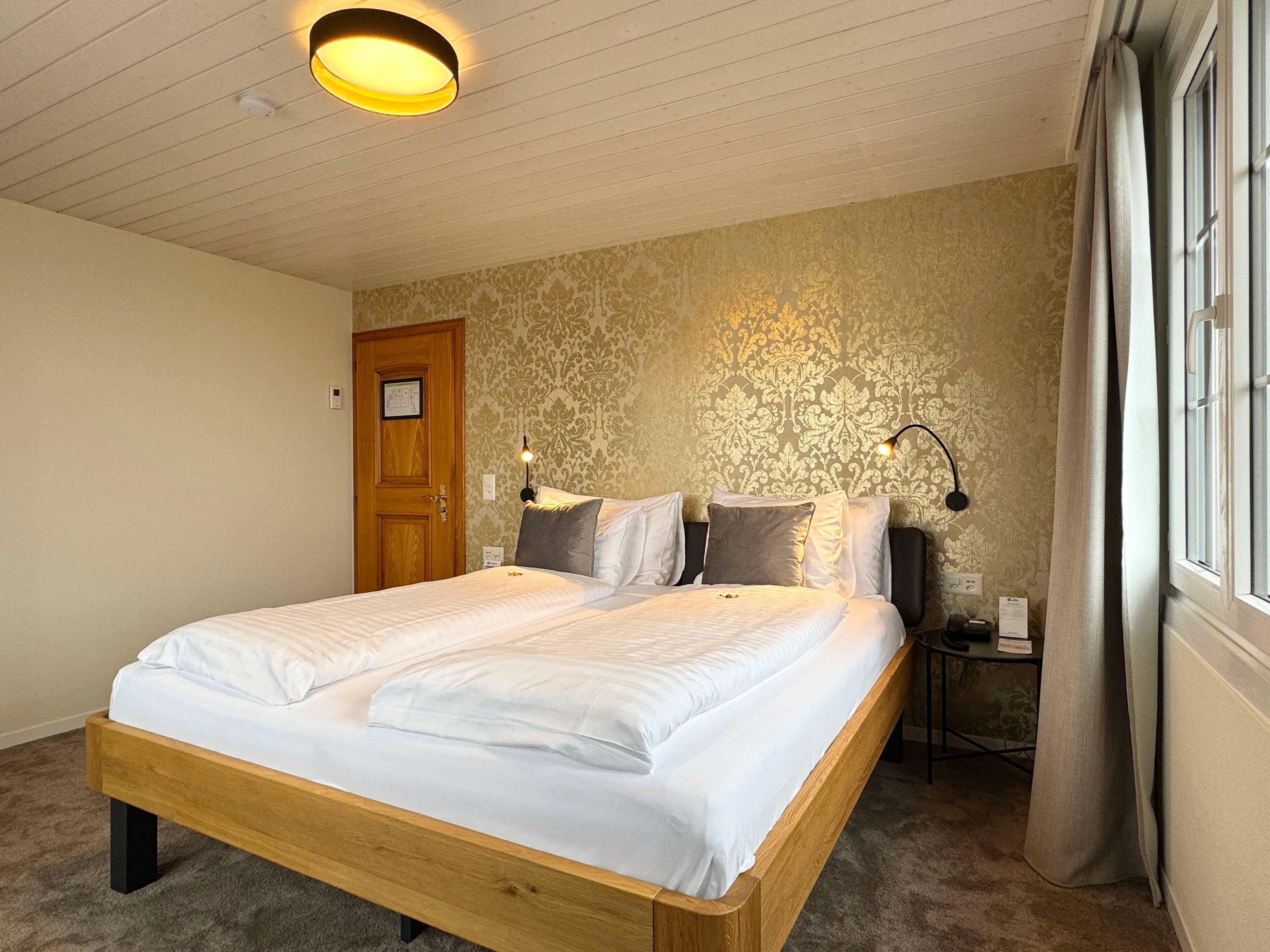 A bedroom with a wooden bed, a nightstand with a lamp, a door, and a patterned wallpaper wall.