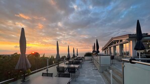 Terraza al aire libre con mesas y sombrillas, con vista a una puesta de sol y al mar.