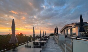 An outdoor terrace with tables and umbrellas, overlooking a sunset and the sea.