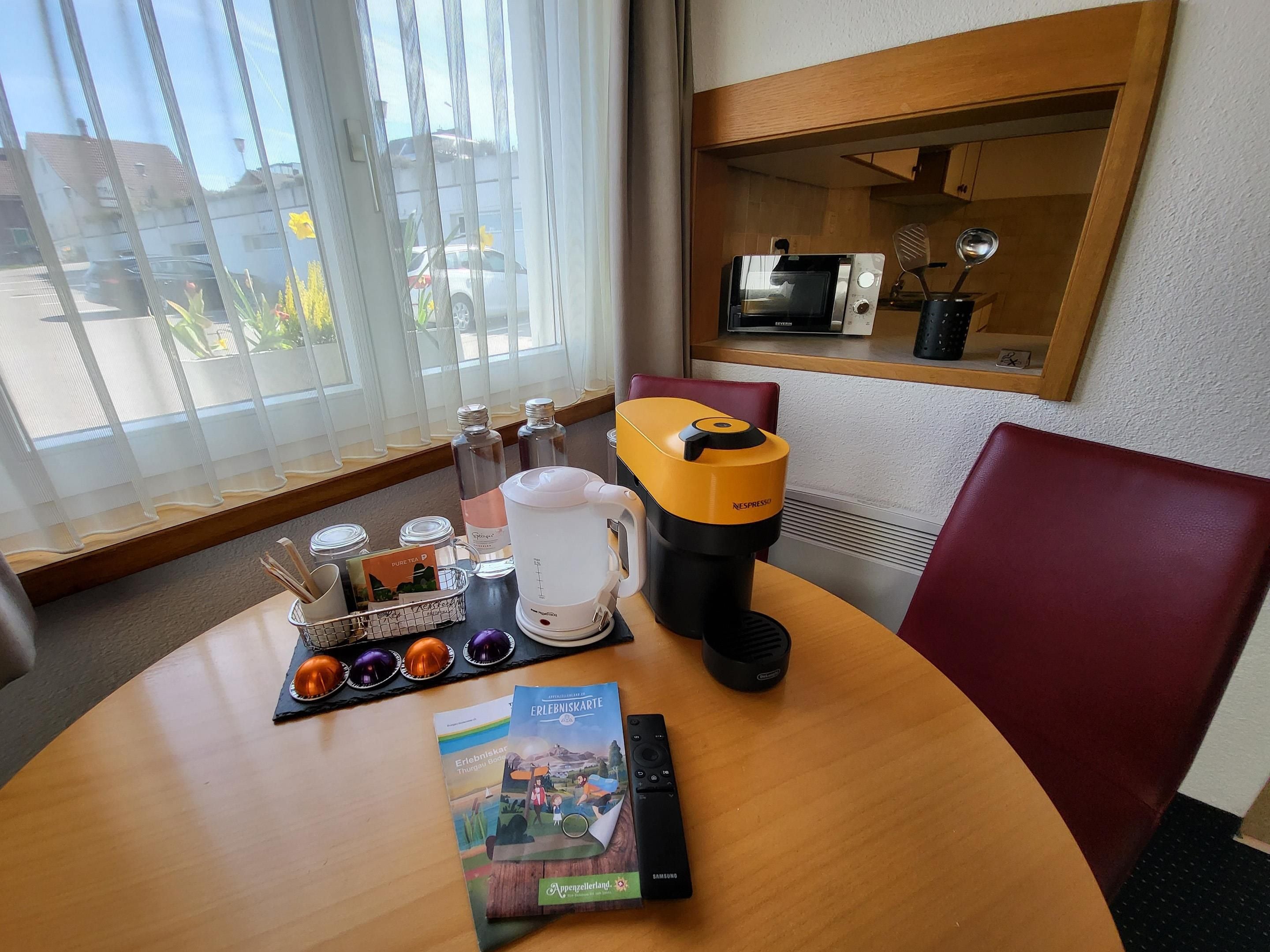 Habitación de hotel con una mesa redonda, una cafetera, un libro y una ventana con vistas al exterior.